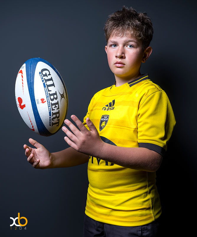 XB1204-6062 Gros plan artistique d’une jeune supportrice du RC Lens, regard vers l'objectif par dessus son épaule avec un ballon noir et or, éclairage studio en contre-jour signé XB1204 Photographe entre Cambrai et Arras.